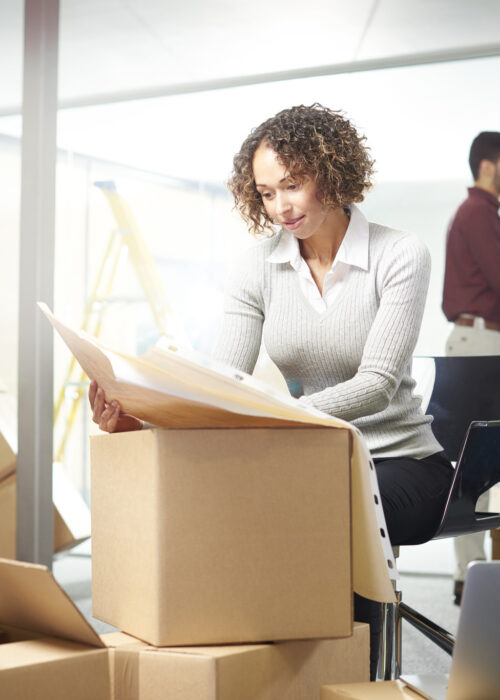 a young female entrepreneur sits at her laptop in an empty office surrounded by moving in boxes but as yet no furniture. Behind her colleagues are organising files, and a male co-worker is chatting to a contractor as to where the furniture needs to be going  . She is looking through the layout of the office on some plans .